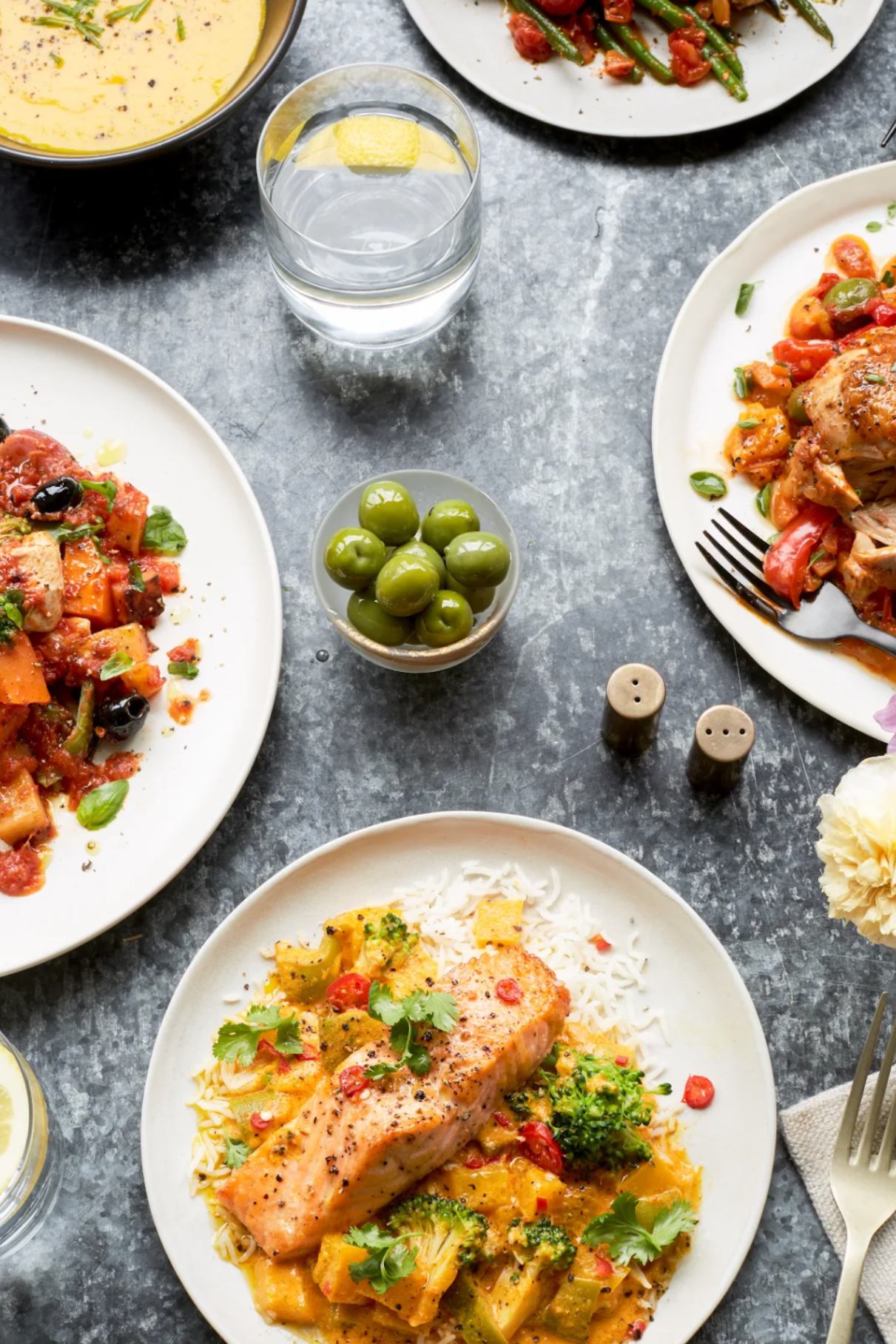 Overhead dining table scene with multiple plated meals, including a salmon dish with yellow sauce and broccoli, a tomato and potato salad, chicken with vegetables, a bowl of green olives, and glasses of water on a gray textured surface.